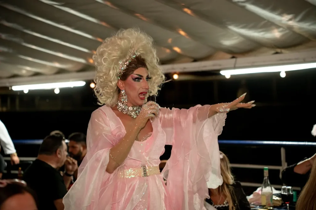Drag queen de rosa cantando con el brazo extendido hacia los invitados bajo la carpa de la cubierta