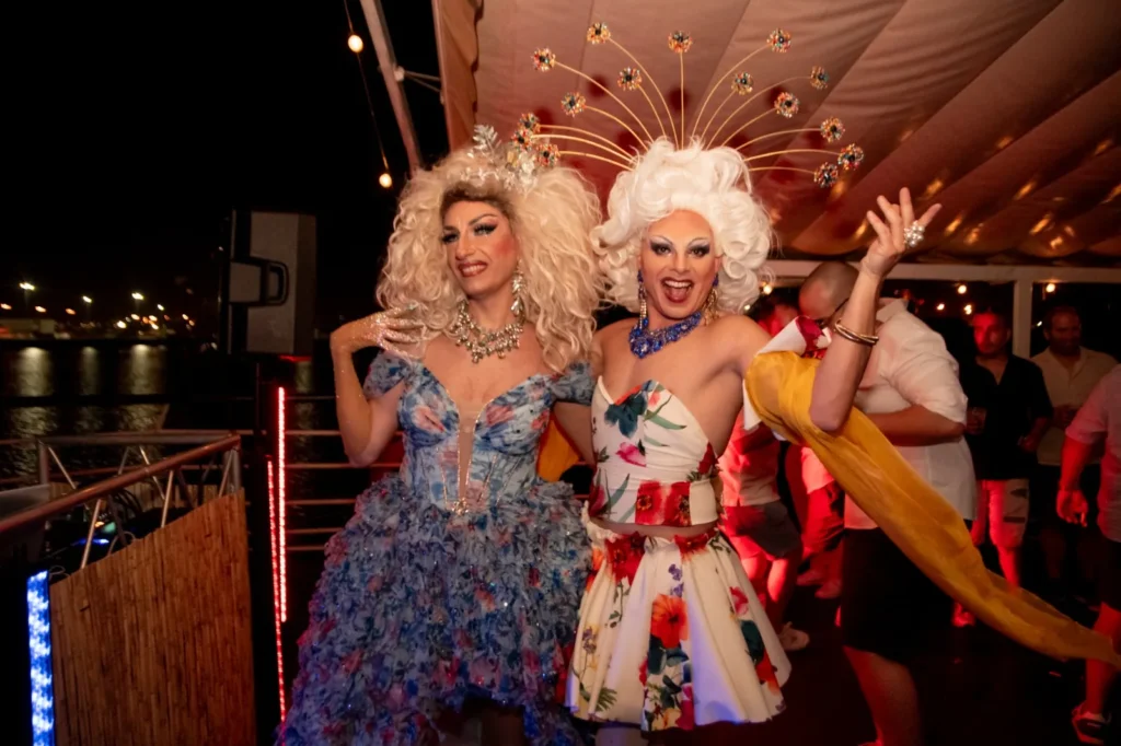 Las drag queens del vestido azul y del vestido de flores posando en la pista del barco bajo la luz roja