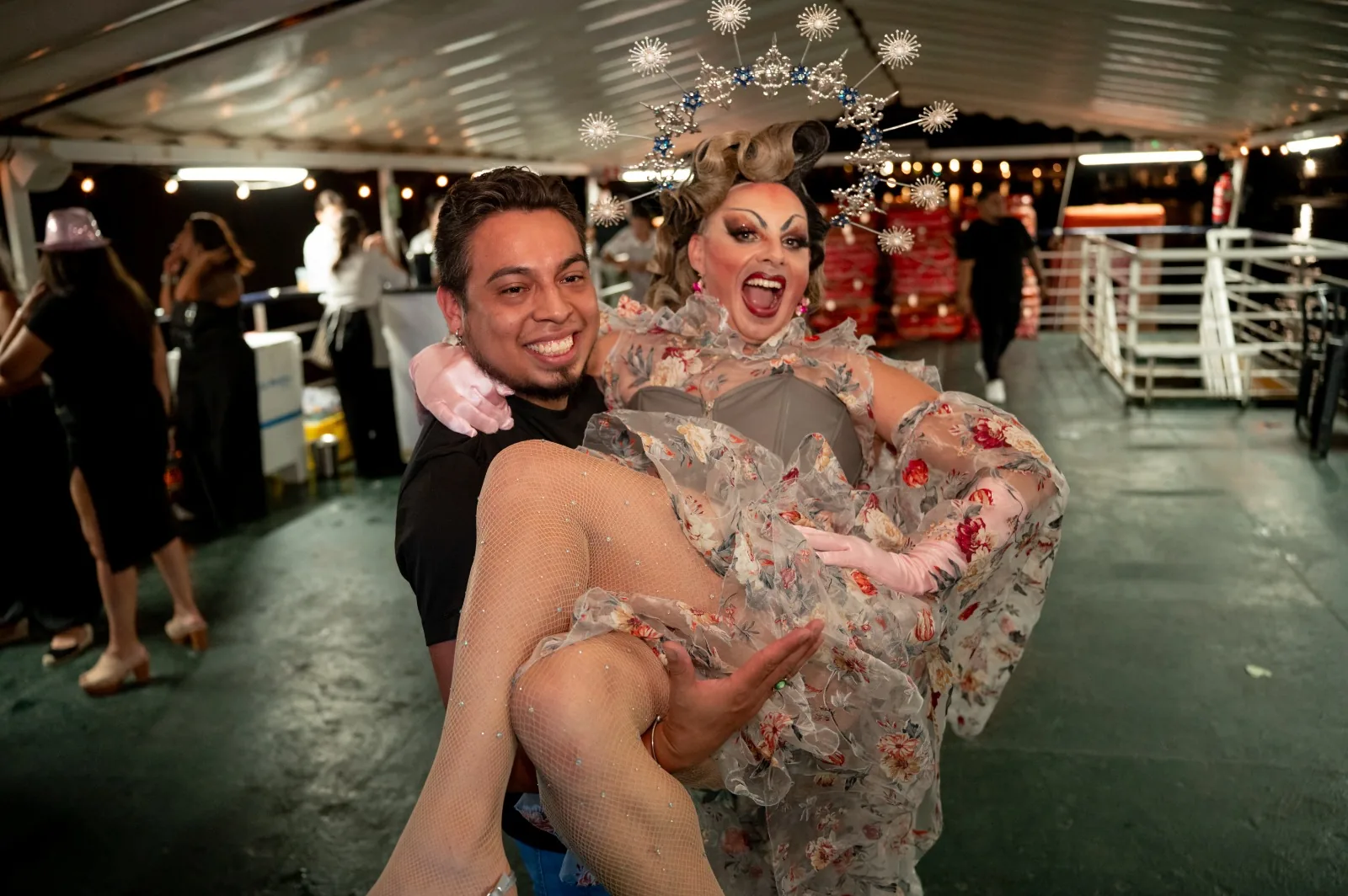 Invitado sonriente llevando en brazos a la drag queen de la corona plateada en la fiesta en barco